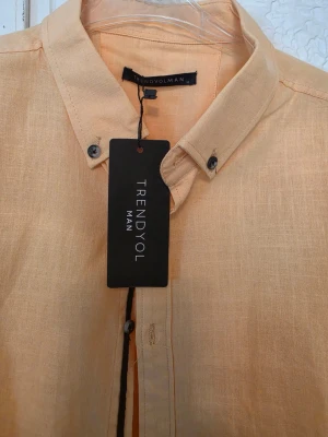 Ljus orange skjorta från Trendyol Man - Snygg ljus orange skjorta från Trendyol Man med klassisk krage och knappar framtill. Skjortan har lång ärm och är tillverkad i ett lätt och luftigt tyg, perfekt för varmare dagar. Enkel och stilren design som passar till många olika outfits.
