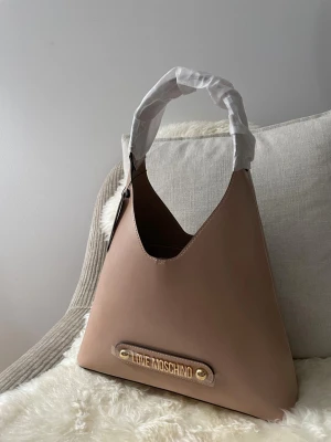 Beige handväska från Love Moschino - Stilren beige handväska från Love Moschino med guldiga detaljer och logotyp framtill. Väskan har en mjuk, triangulär form och ett brett axelband. Tillverkad i slätt konstläder som ger en lyxig känsla.