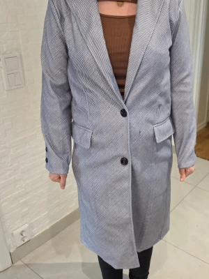 Ljusgrå lång kappa med smala ränder - Snygg ljusgrå kappa med smala vita ränder och klassisk krage. Kappan har tre svarta knappar framtill, fickor med lock och dekorativa knappar vid ärmsluten. Rak passform och stilren look som passar till många outfits.