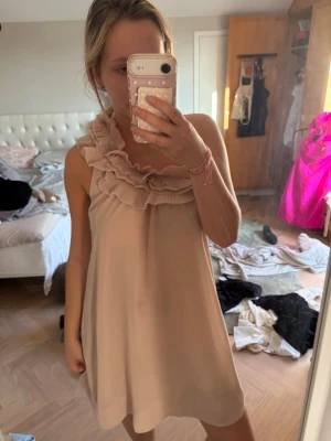 Beige kort klänning med volang - Supersöt populär beige kort klänning med volangdetaljer runt halsringningen. Klänningen är ärmlös och har en lös passform som ger en avslappnad och trendig look. Perfekt för varma dagar och när du vill känna dig extra fin. 
