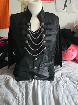 Svart militärinspirerad cosplayjacka - Snygg svart cosplayjacka med militärstil, dekorerad med silverfärgade kedjor och knappar framtill. Jackan har ståkrage, långa ärmar och detaljer i både tyg och fuskläder. Perfekt för maskerad eller cosplay med en edgy look. Jag upskattar storleken till XL, förra ägaren hade klippt av lapparna.