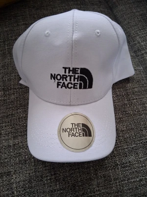Vit keps från The North Face - Snygg vit keps från The North Face med broderad logga i svart framtill. Klassisk böjd skärm och justerbar baksida. Tillverkad i slitstarkt tyg med ventilerande hål upptill för extra komfort. Perfekt accessoar för en sportig och trendig look.