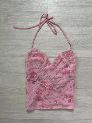 Rosa broderad halterneck topp/ Pink sequin glitter top - Supersöt rosa halterneck topp med broderade blommor och paljettdetaljer. Toppen har snörning i ryggen och är croppad, perfekt för en trendig look. Materialet känns lätt och toppen har en feminin v-ringning. DEN ÄR XXS MEN PASSAR MIG JÄTTE BRA SOM S! OCH ÄR HELT NY, ANVÄNT EN GÅNG! Den är slutsåld i butiken!