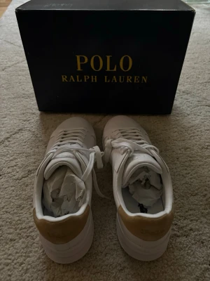 Vita Polo Ralph Lauren sneakers (nya) - Snygga vita sneakers från Polo Ralph Lauren med klassisk låg profil. Skorna har vita snören, gulddetaljer vid hälen och Polo-loggan broderad på sidan. Tillverkade i slätt läder för en clean och stilren look.