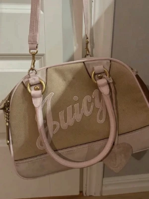 Beige och rosa Juicy handväska - Snygg handväska från Juicy Couture i beige canvas med ljusrosa detaljer i mocka och guldiga metalldelar. Väskan har både axelrem och handtag, samt dekorativa kedjor och ett stort Juicy-tryck på framsidan. Perfekt för dig som gillar Y2K-stil!