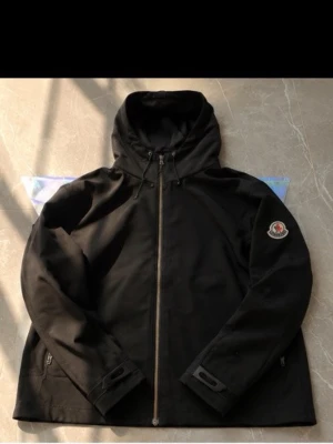 Svart vindjacka från Moncler - Stilren svart vindjacka från Moncler med huva och hel dragkedja framtill. Jackan har två sidofickor med dragkedja och klassisk Moncler-logga på vänster ärm. Perfekt för blåsiga dagar och enkel att matcha med olika outfits.