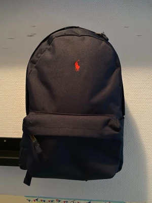 Mörkblå ryggsäck från Polo Ralph Lauren - Snygg mörkblå ryggsäck från Polo Ralph Lauren med klassisk röd logga framtill. Ryggsäcken har ett stort huvudfack, en framficka med dragkedja och justerbara axelband. Tillverkad i slitstarkt tyg, perfekt för skolan eller stan.
