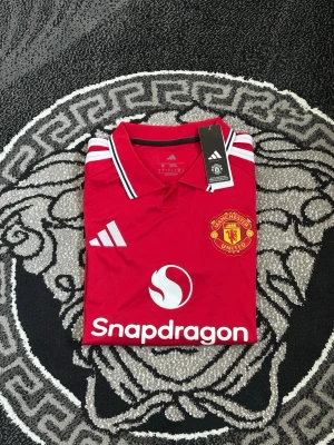Manchester United matchtröja röd - Snygg röd Manchester United matchtröja från Adidas med klubbmärke på bröstet och vita detaljer. Tröjan har vit logga, sponsortryck och klassiska Adidas-ränder på axlarna. Tillverkad i lätt och ventilerande material, perfekt för fotboll eller träning.