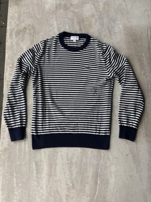 Randig stickad tröja från Lacoste - Snygg stickad tröja från Lacoste med marinblå och vita horisontella ränder. Perfekt nu när randigt är trändigt och känslan av tröjan skriker stilren!