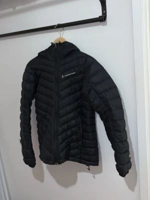 Svart peak performance - Snygg svart dunjacka från Peak Performance med huva och dragkedja framtill. Jackan har ett quiltat mönster i sicksack, två sidofickor med dragkedja och logga på bröstet. Perfekt för kalla vinterdagar och riktigt lätt att bära, men den har tyvärr ett håll i sig.