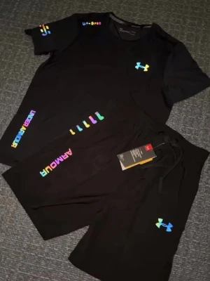 Svart träningsset från Under Armour - Snyggt svart träningsset från Under Armour med färgglada loggor och detaljer. Setet består av en t-shirt och shorts, båda med UA-loggan i regnbågsfärger. Perfekt för gymmet eller löprundan, tillverkad i ett lätt och snabbtorkande material.