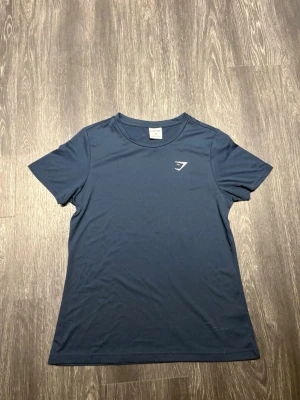 Blå t-shirt från Gymshark - Gymshark Training T-Shirt i mörk marinblå färg, storlek M. Stilren och minimalistisk design med klassisk Gymshark-logga på bröstet. Tillverkad i lätt, mjukt och stretchigt funktionsmaterial med atletisk passform och bra rörelsefrihet. En mångsidig träningströja som passar perfekt för gym, löpning och vardag