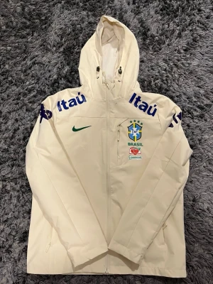 Vit Brasil Nike vindjacka med huva - Snygg vit vindjacka från Nike med Brasiliens landslagslogga och sponsorer tryckta på bröstet och ärmarna. Jackan har huva med dragsko, hel dragkedja framtill och två sidofickor. Perfekt för dig som gillar sportig stil och vill sticka ut.