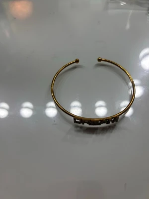 Guldfärgat armband med text DREAM - Snyggt och stilrent armband i guldfärg med öppen design och texten 'DREAM' framtill. Bokstäverna är dekorerade med små glittrande stenar som ger extra bling. Perfekt accessoar för dig som gillar statement-smycken och vill ha något unikt☺️ (aldrig använt)