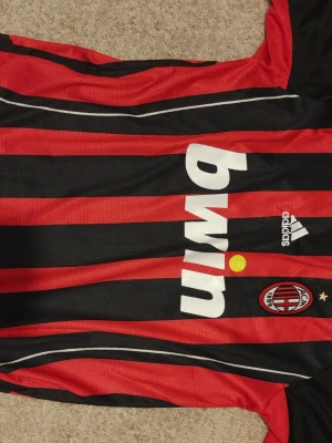 AC Milan Adidas matchtröja röd/svart - AC Milan matchtröja från Adidas med klassiska röda och svarta vertikala ränder, klubbmärke på bröstet och Bwin som sponsor. Tröjan har korta ärmar och är gjord i ett lätt, ventilerande material som passar perfekt till fotbollsträning.