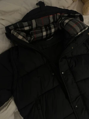 Svart pufferjacka med rutigt foder - Säljer en svart pufferjacka från Burberry med klassiskt rutigt foder i huvan. Jackan har dragkedja, tryckknappar och två fickor framtill. På ärmen finns Burberry London England-tryck. Perfekt för kalla vinterdagar.