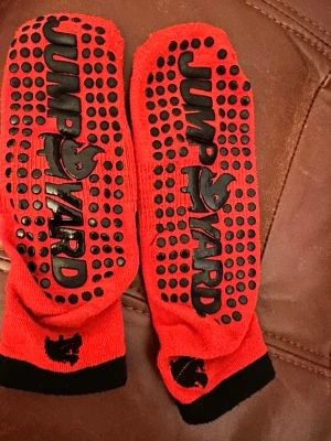 Röda JumpYard halksockor 20-25cm längd fot  - Säljer ett par röda halksockor från JumpYard med svarta detaljer och logga. Strumporna har svarta halkpluppar undertill för extra grepp och svart kant vid ankeln. Perfekta för trampolinpark eller inomhuslek.