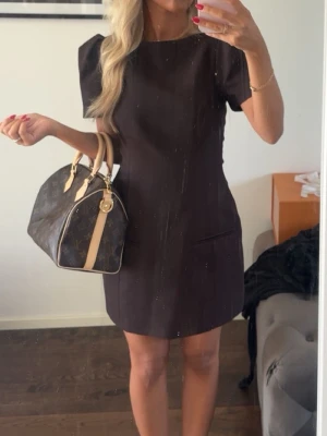 Mörkbrun kort klänning med puffärm - Snygg mörkbrun kort klänning med puffiga axlar och raka linjer. Klänningen har korta ärmar och två fickor framtill. Perfekt för dig som gillar en stilren och modern look.