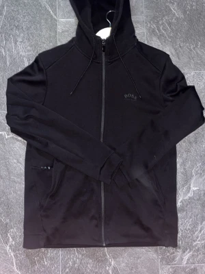 Svart zip hoodie från BOSS - Svart zip hoodie från Hugo boss. Den har ett tjockare exklusivt material på ryggen samt vanligt framtill. Väldigt lite använd. Väldigt bra passform, jag är 182 och ni kan se hur den sitter på bilderna. Storlek S men passar även M. Perfekt nu till höst/vinter för olika typer av använding. Pris kan diskuteras, skriv vid frågor!