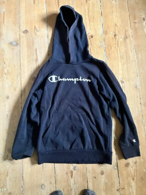 Svart hoodie från Champion L - Svart hoodie från Champion med stor logga över bröstet och en klassisk känguruficka framtill. Tröjan har huva och ribbade muddar vid ärmslut och nederkant. Perfekt för en avslappnad och sportig stil.