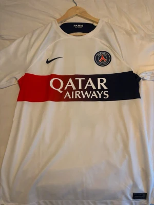 PSG 23/24 borta kit MBAPPE  - Snygg vit PSG matchtröja från Nike med Mbappé och nummer 7 på ryggen. Tröjan har klubbmärke på bröstet, svart Nike-logga och breda ränder i rött och marinblått över bröstet. Tillverkad i lätt och ventilerande material, perfekt för fotboll eller träning.
