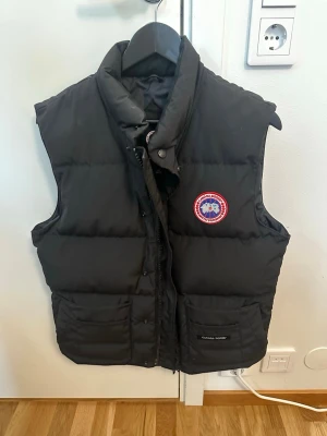 Svart dunväst från Canada Goose - Snygg svart dunväst från Canada Goose med klassisk logga på bröstet. Västen har hög krage, två stora fickor framtill och stängs med dragkedja och knappar. Perfekt för lager-på-lager och håller dig varm under kyliga dagar.