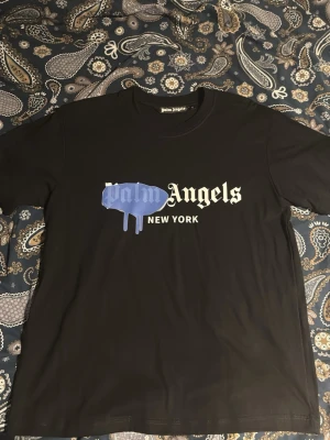 Palm Angels svart t-shirt New York - Svart t-shirt från Palm Angels med tryck i vitt och blått framtill där det står 'Palm Angels New York' och en blå drip-effekt över texten. Klassisk rund hals och korta ärmar. Perfekt för dig som gillar streetwear och vill sticka ut.