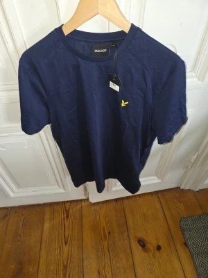 Mörkblå t-shirt från Lyle & Scott - Snygg mörkblå t-shirt från Lyle & Scott med klassisk rund hals och korta ärmar. T-shirten har en gul broderad logga på bröstet och är tillverkad i mjuk bomull. Perfekt för en stilren och enkel look.