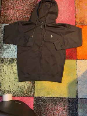 Svart hoodie från Polo Ralph Lauren - Snygg svart hoodie från Polo Ralph Lauren med dragkedja, huva med dragsko och klassisk vit logga broderad på bröstet. Tillverkad i mjukt material och har två fickor framtill. Perfekt för en chill och stilren look.