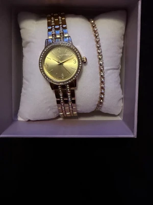 Guld klocka med strass & diamant armband från Regal Ur & Penn  - Snygg guldfärgad klocka från Regal med glittrande strass runt urtavlan och på medföljande armband. Klockan har en klassisk rund urtavla i guld och matchas med ett elegant armband med stenar/diamanter. Passar också perfekt som present.