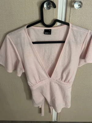 Ljusrosa omlottopp från Gina Tricot - Supersöt ljusrosa omlottopp från Gina Tricot med korta ärmar och knyt i midjan. Toppen har en djup v-ringning och är gjord i mjuk bomull, perfekt för en trendig och avslappnad look.