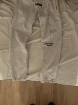 Gråa Essentials mjukisbyxor - Säljer ett par gråa mjukisbyxor från Fear of God Essentials. Byxorna har resår i midjan och benslut, dragsko och tryckt logga på benet. Tillverkade i mjukt och skönt material som passar perfekt för chill dagar.