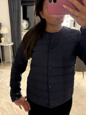 Uniqlo jacka - Snygg mörkblå pufferjacka med quiltad design och knappar framtill. Jackan har en enkel, stilren look utan krage och långa ärmar. Perfekt för kyligare dagar och lätt att matcha med olika outfits.
