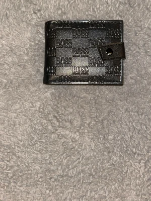 Svart plånbok - Snygg svart plånbok  i läder med präglad logga över hela ytan. Plånboken har en praktisk knappstängning och en klassisk, fyrkantig form. Perfekt för dig som vill ha en stilren accessoar med tydlig logotyp.