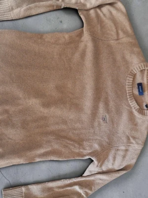 Beige stickad tröja från Gant - Snygg beige stickad tröja från Gant med rund halsringning och diskret broderad logga på bröstet. Tröjan har långa ärmar och ribbade muddar vid ärmslut och nederkant. Perfekt för lager på lager och enkel att matcha.