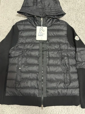 Svart dunväst med huva från Moncler - Snygg svart dunväst från Moncler med huva och ribbade ärmar. Västen har två sidofickor med dragkedja och en Moncler-logga på ärmen. Tillverkad i glansigt material med quiltad design och dragkedja framtill. Perfekt för lager-på-lager och streetstyle.