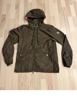 Olivgrön vindjacka från Moncler med huva - Snygg olivgrön vindjacka från Moncler med dragkedja framtill, två fickor med tryckknappar och justerbar huva med snören. Jackan har Moncler-logga på ärmen och är tillverkad i ett lätt, vindtätt material. Perfekt för blåsiga dagar.