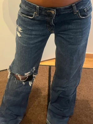 Baggy bootcut jeans  - Säljer ett par mörkblå bootcut jeans med slitningar och hål på vänster lår och knä. Jeansen har klassisk femficksmodell, låg midja och råa kanter vid hålen. Perfekta för en avslappnad och trendig look.