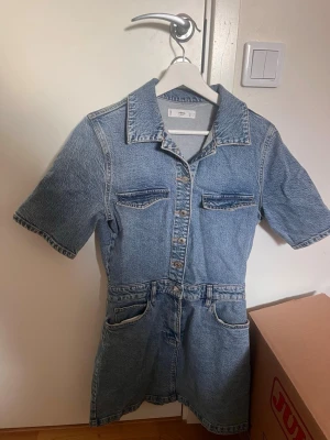 Kort jeansklänning från MNG Denim - Kort jeansklänning från MNG Denim med skjortkrage, korta ärmar och knappar framtill. Klänningen har fickor både upptill och nedtill och är gjord i klassiskt blått denimtyg. Enkel och trendig modell.