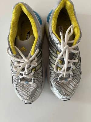Grå och gula Adidas sneakers - Snygga sneakers från Adidas i grått med gula och blå detaljer. Ovandelen är i mesh och syntet för bra andningsförmåga. Klassiska tre ränder på sidan och vit sula. Skön innersula och vadderad kant i gult. Perfekta för en sportig look.