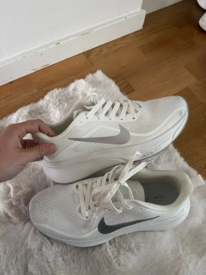 Vita Nike sneakers med silverdetaljer - Snygga vita Nike sneakers med silverfärgad swoosh på sidan. Skorna har en luftig mesh-överdel, vita skosnören och en tjock, bekväm sula. Perfekta för dig som gillar stilrena och sportiga sneakers med modern design.