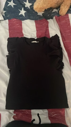 Svart topp med volangdetaljer - Säljer en svart topp med volangdetaljer längs axlarna. Toppen har en enkel, stilren design och är perfekt för dig som gillar en clean look med lite extra edge. Passar till det mesta och är lätt att matcha med olika outfits.
