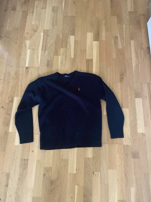 Mörkblå stickad tröja Polo Ralph Lauren STORLEK L - Klassisk mörkblå stickad tröja från Polo Ralph Lauren med V-ringning och den ikoniska röda logotypen broderad på bröstet. Tröjan har ribbstickade muddar vid ärmslut och nederkant. Perfekt för lager på lager och stilrena outfits.
