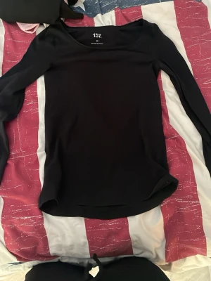Svart långärmad topp från 157 - En stilren svart långärmad topp från 157 med rund halsringning. Toppen har en enkel och klassisk design som passar till många olika outfits. Materialet känns mjukt och stretchigt, perfekt för en bekväm vardagslook.