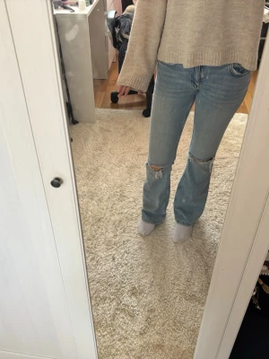 Ljusblå bootcut jeans med hål - Säljer ett par ljusblå bootcut jeans med slitna hål på båda knäna. Jeansen har klassisk femficksdesign och är tillverkade i denim. Perfekta för en avslappnad och trendig look.