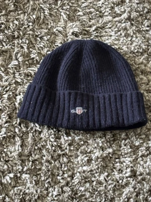 Mörkblå ribbad mössa från Gant - Säljer en mörkblå ribbstickad mössa från Gant med uppvikt kant och broderad logga framtill. Klassisk och enkel design som passar perfekt till kyliga dagar. Materialet känns mjukt och värmande.