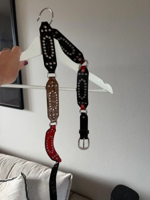 Svart, brunt och rött bälte med nitar - Unikt bälte i svart, brunt och rött med silvriga nitar och ringdetaljer. Bältet har breda partier och mixar slätt och krokopräglat skinn. Perfekt för att ge outfiten en edgy vibe och sticker verkligen ut med sina kontrasterande färger och detaljer.