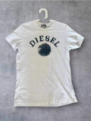 Vit t-shirt från Diesel med tryck - Säljer en vit t-shirt från Diesel med ett stort svart tryck och texten DIESEL på bröstet. T-shirten har klassisk passform, rund halsringning och korta ärmar. Perfekt till jeans eller shorts för en avslappnad look.
