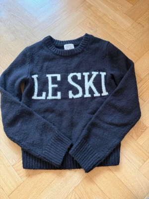 Svart stickad tröja LE SKI - Mysig svart stickad tröja med vit text 'LE SKI' framtill. Tröjan har rund halsringning, långa ärmar och ribbade muddar vid ärmslut och nederkant. Perfekt för kalla dagar och en cool tröja till en skiddag i Alperna 🫶🏼🫶🏼