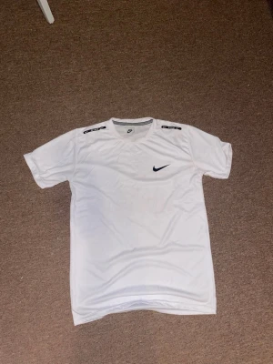 Vit Nike t-shirt med logga - Säljer en vit t-shirt  med svart logga på bröstet och svarta Nike-detaljer på axlarna. Klassisk rund hals och korta ärmar. Perfekt för dig som gillar sportig och enkel stil.